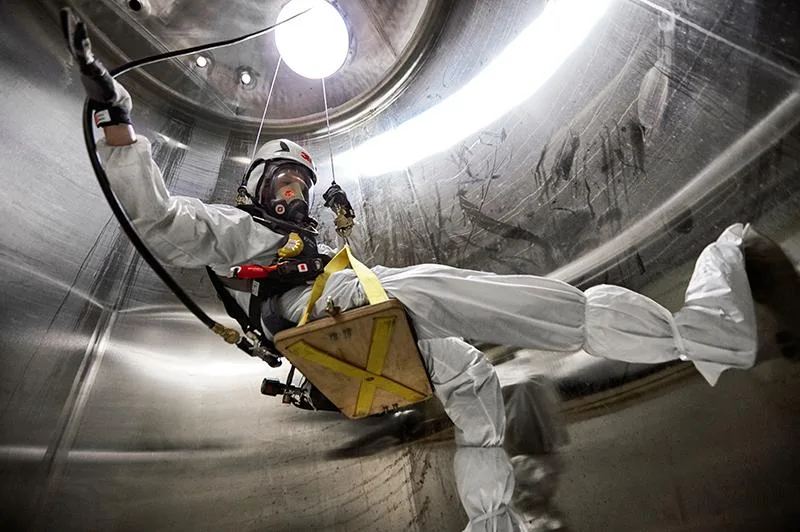 A worker in protective overalls and a breathing mask inside a large steel tank