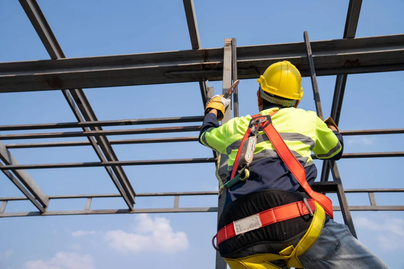 A worker in a hard hat and fall-arrest harness climbing a steel structure against the sky